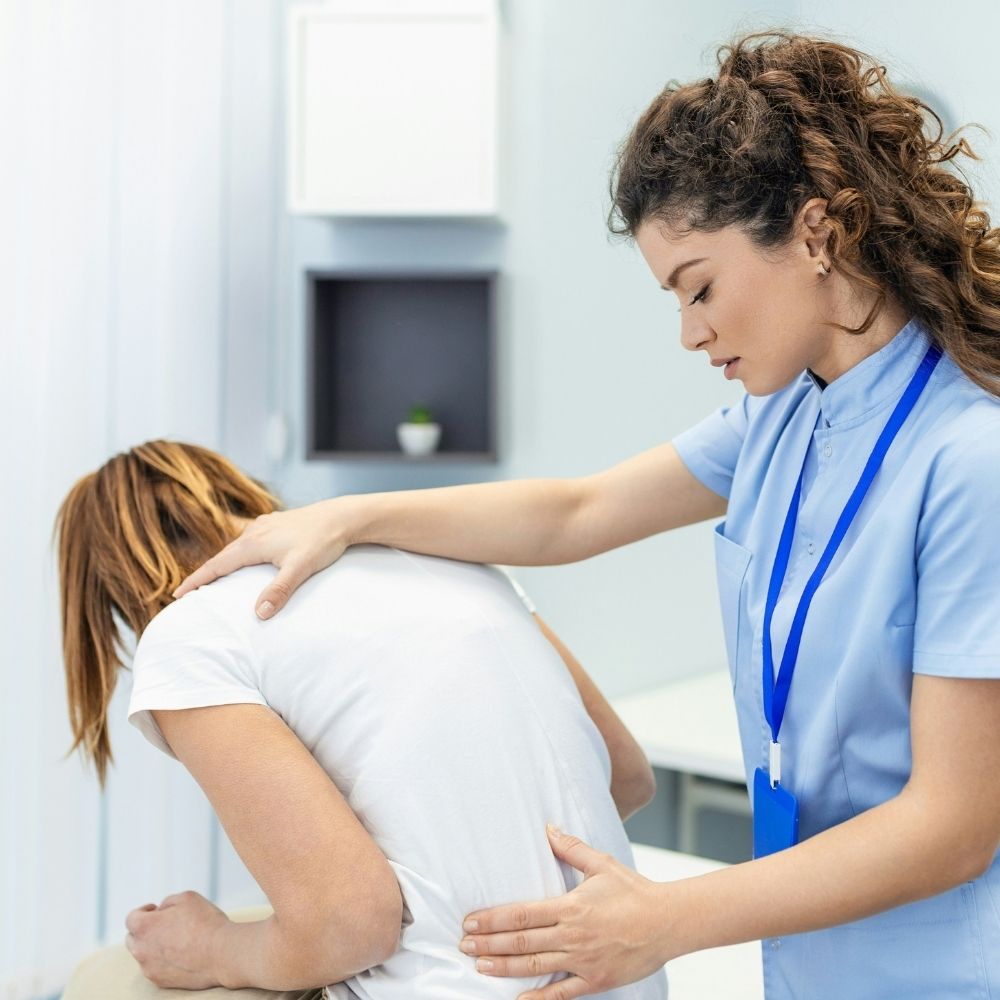 ASA (13) A physical therapist in blue scrubs examines a patients back while the patient sits hunched forward on an exam table.