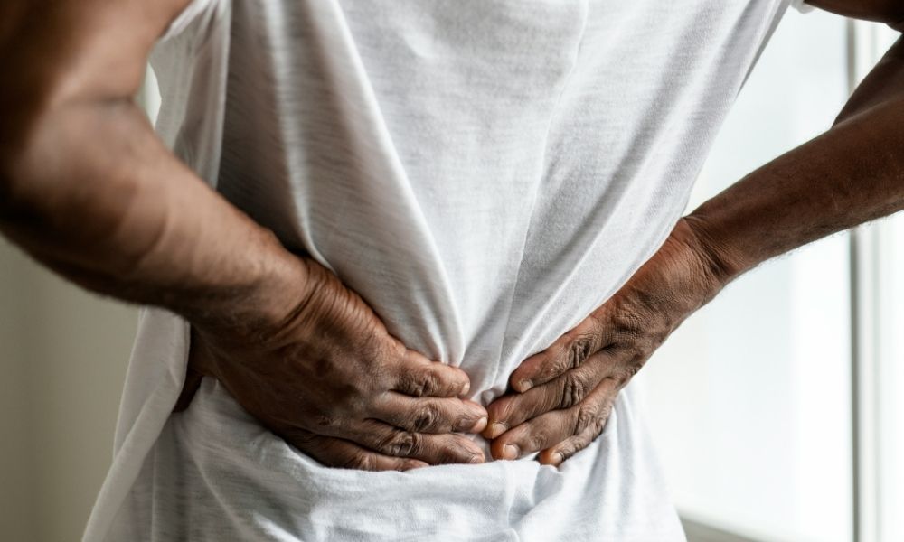 A person in a white shirt holding their lower back with both hands, indicating pain.
