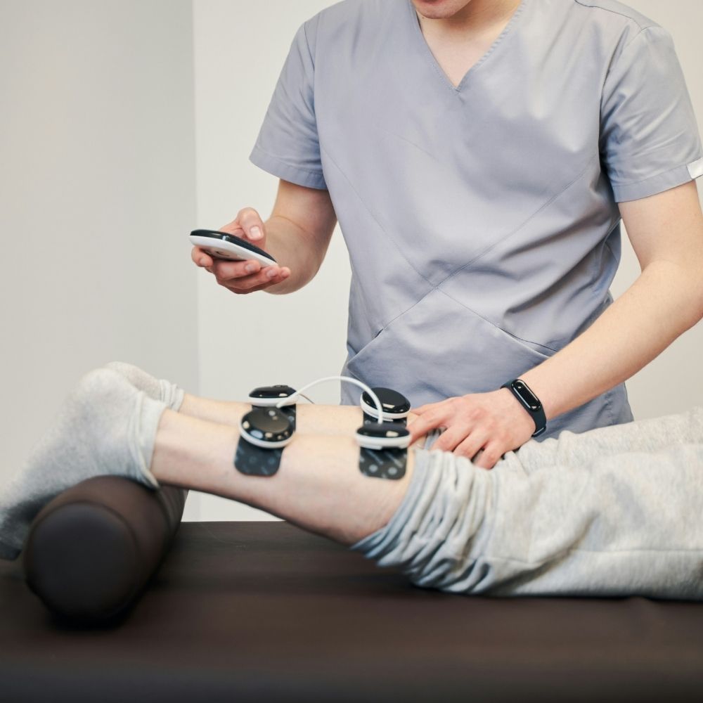 A therapist adjusts electronic muscle stimulation pads on a patients lower legs while holding a remote control.