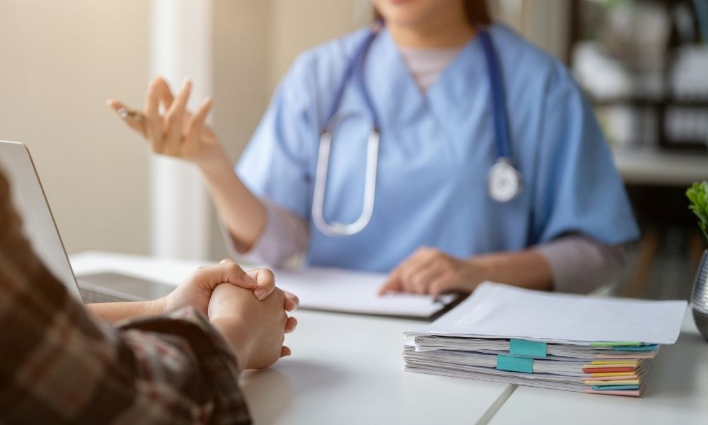 A healthcare professional in blue scrubs with a stethoscope around their neck talks to a patient across a desk with documents.
