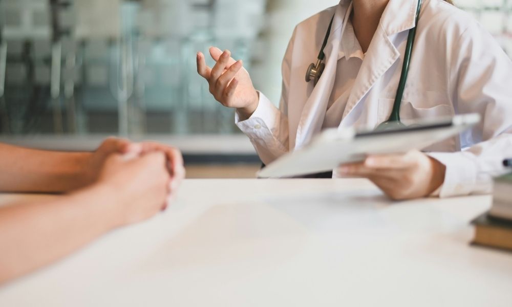 A doctor in a white coat and stethoscope talks to a patient across a table.