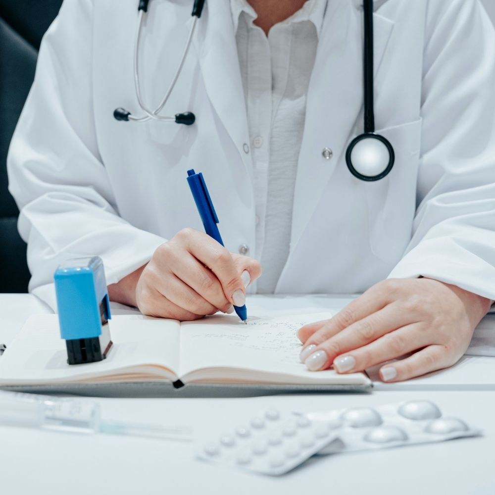 A doctor in a white coat with a stethoscope around their neck writes in a notebook, with a stamp and blister packs of pills on the desk.