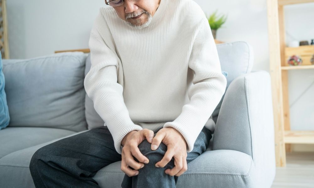 An older man sitting on a sofa and holding his knee in pain.