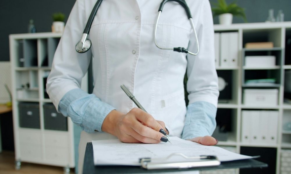A doctor in a white coat with a stethoscope around their neck writes on a clipboard.
