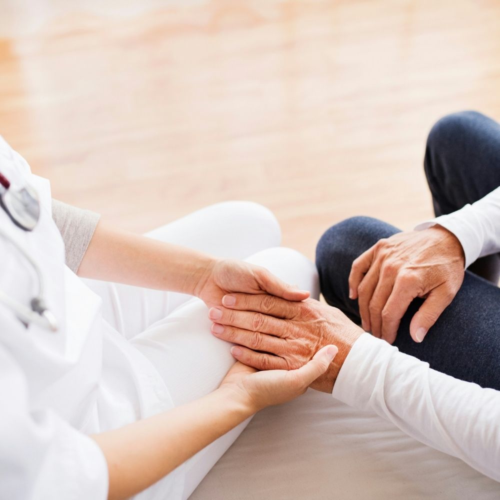 A healthcare worker in white scrubs holds the hands of an older patient.
