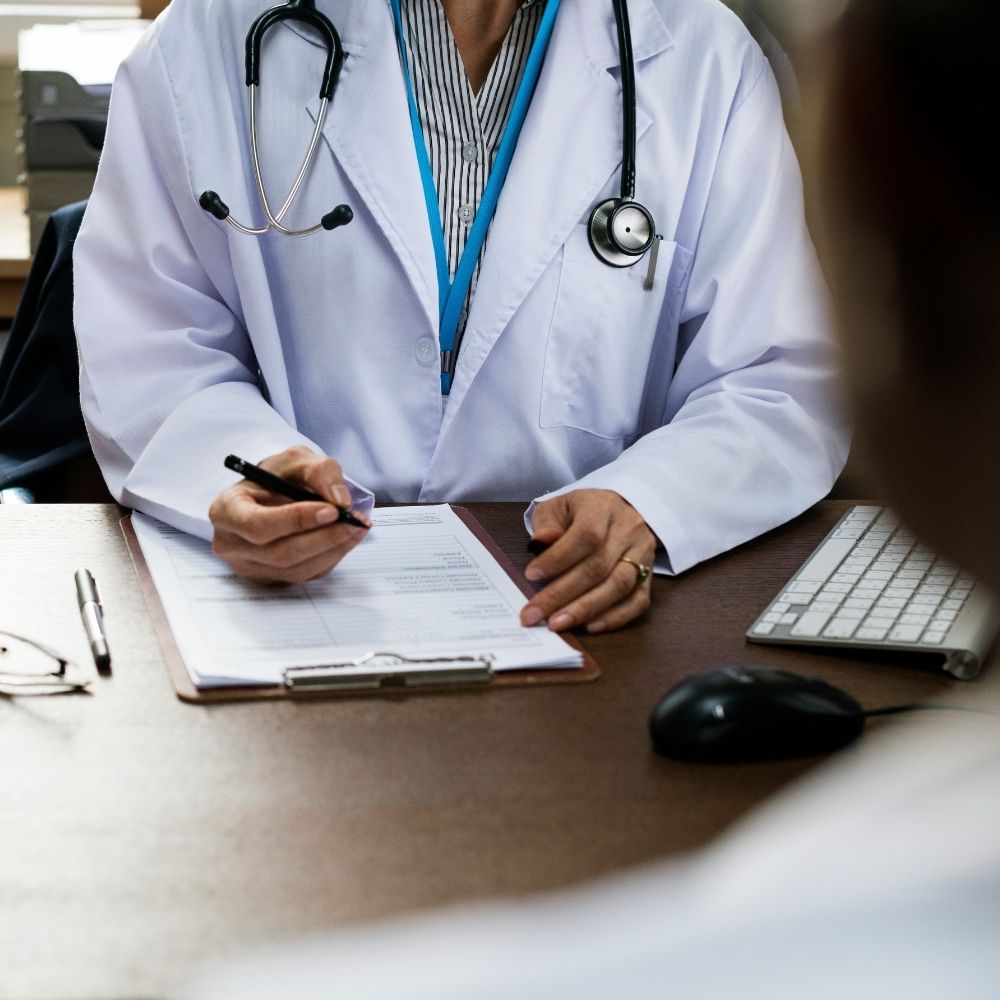 A doctor in a white coat with a stethoscope around their neck writing on a clipboard at a desk.