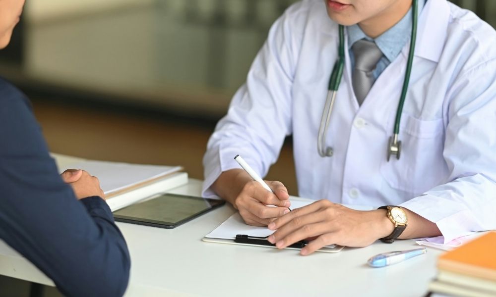 A doctor in a white coat and stethoscope writes on a clipboard while talking to a patient across a desk.