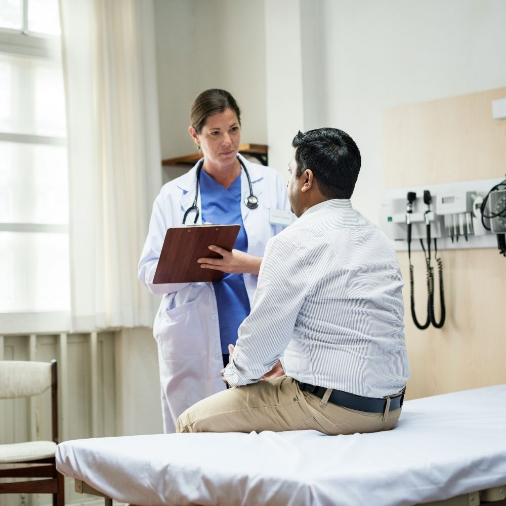 A female doctor in a white coat with a stethoscope holds a clipboard while speaking to a male patient sitting on an exam table.