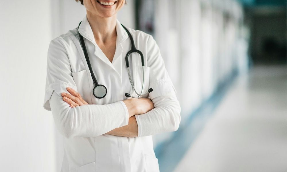 A smiling doctor in a white coat with a stethoscope around her neck stands with her arms crossed in a hospital hallway.