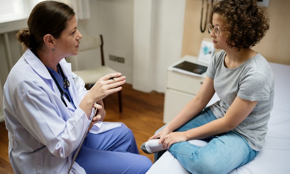 A doctor in a white coat talks to a young patient sitting on an exam table.