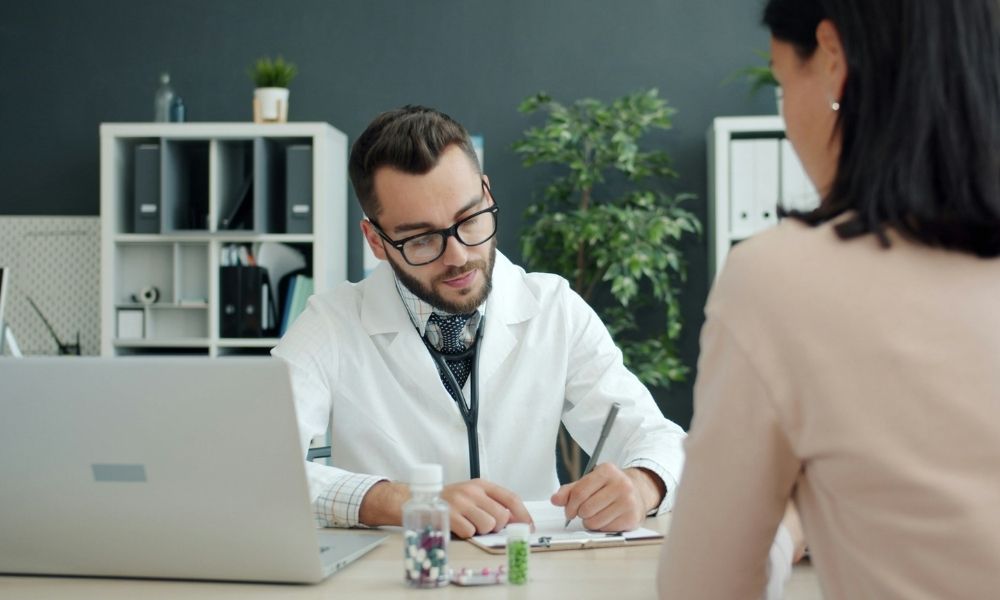 A doctor in a white coat writes on a notepad during a consultation with a patient in an office.